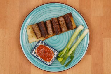 Yeşil soğanlı ve mavi tabakta ayvar bulunan bir ekmek üzerinde doğranmış et parmakları (cevapi).
