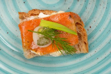 Vejetaryen füme somonu andırmaya hazırlanan havuç dilimli küçük bir sandviç krem peynir, dereotu, soğan ve kapari açık mavi ve kahverengi benekli bir tabağın üzerinde oturur.