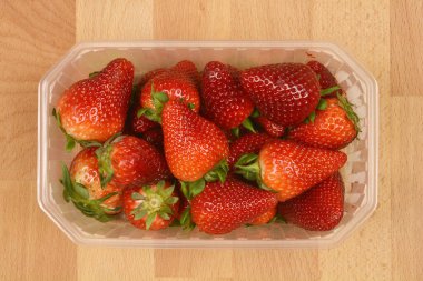 Açık bir plastik kap, açık kahverengi ahşap bir yüzeye yerleştirilmiş yeşil saplı olgun kırmızı çilekleri barındırır.