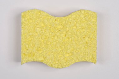 Dalgalı üst ve alt kenarlı açık sarı bir sünger düz açık gri bir arkaplanda merkezlenir.