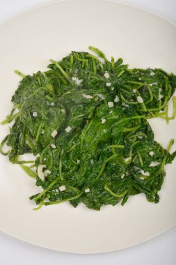 Tepetaklak bir perspektiften beyaz bir tabakta görünen sarımsak parçalarıyla pişirilmiş turp yaprakları sunuluyor.