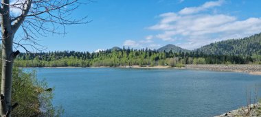 Geniş bir açı Gorski Kotar 'daki sakin Lepenica Gölü' nü yakalıyor. Etrafı yemyeşil ormanlar ve dağınık mavi bulutlarla kaplı uzak dağlarla çevrili.