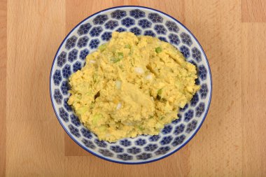 Yukarıdan aşağıya yakın plan mavi ve beyaz desenli bir kase gösteriyor. İçi kremalı, açık sarı karışımlı yumurta püresi, avokado ve doğranmış soğan dolu. Baharatlı yumurtalar için hazırlanmış, ahşap bir yüzey.