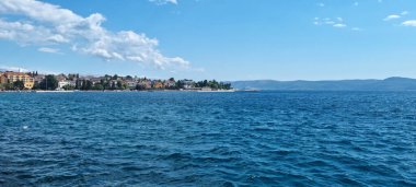 Hırvatistan 'ın Selce kentinin pitoresk kıyı manzarası, açık mavi Adriyatik Denizi boyunca parçalı bulutlu bir gökyüzünün altında uzanan renkli binaları gösteriyor