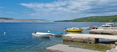 Jadranovo 'daki pitoresk bir limanda beton rıhtımlara demirlemiş çok sayıda küçük tekne bulunur ve Krk Köprüsü mavi denizin üzerinde parçalı bulutlu bir gökyüzünün altında görülebilir.