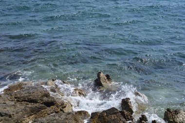 Koyu mavi deniz dalgaları engebeli kayalara çarpar. Kıyı boyunca uzanan koyu kayalar doğal ışık altında beyaz köpüklü sıçramalar yaratır.