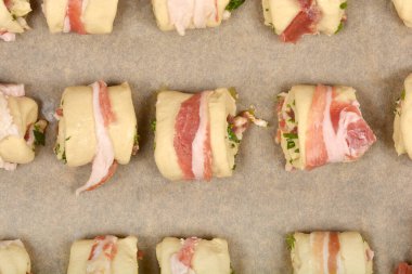 Pastırmaya sarılmış hamur ruloları dilimlenmiş pancetta karışımı ve otlar pişirilmeye hazır bir tepside dizilmiştir.