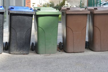 Farklı renklerde kapaklı tekerlekli çöp bidonları asfalt bir yüzeye oturur.