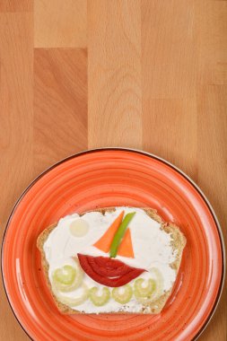 Yaratıcı bir şekilde dekore edilmiş krem peynirli ve sebzeli sandviç kırmızı bir tabakta, dalgalar üzerindeki bir tekneye benziyor.