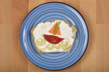 Dalgalardaki bir tekneye benzeyen mavi tabakta krem peynirli ve sebzeli sandviç.