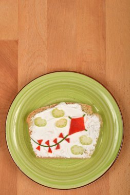 Bu resim, krem peynirle kaplanmış, uçurtma ve bulut şeklinde sebze parçalarıyla süslenmiş, ahşap bir yüzeyde yeşil bir tabakta servis edilen bir ekmek dilimini gösteriyor.