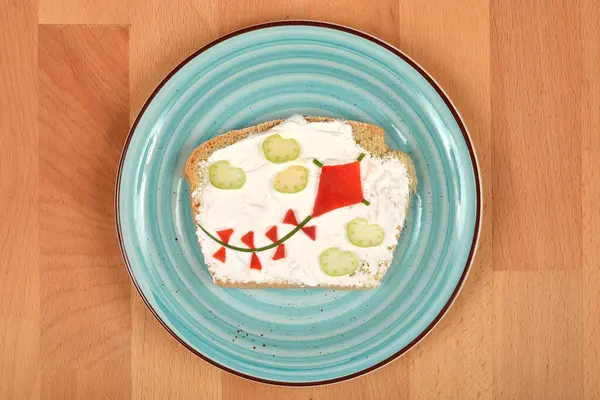 Bu resim, krem peynirle yayılmış, uçurtma ve bulut şeklinde sebze parçalarıyla süslenmiş, ahşap bir yüzey üzerinde mavi bir tabakta servis edilen bir ekmek dilimini gösteriyor.