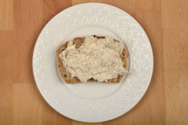 Yukardan aşağı doğru çekimde, üzerinde kremalı ton balığı olan bir parça ekmek, merkezde beyaz desenli bir tabakta, ahşap bir yüzey üzerinde yer alır.