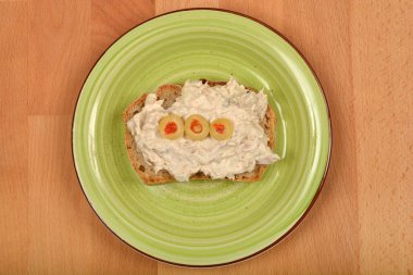 Yukarıdan aşağıya doğru kaymak gibi bir ton balığıyla kaplanmış ve merkezde açık yeşil spiral desenli bir tabağa yerleştirilmiş üç yeşil zeytinle süslenmiş bir ekmek parçası.