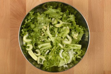 Tepeden çekimde metal bir kasede taze yeşil bir salata, yapraklı yeşiller ve dilimlenmiş kereviz sergileniyor, ahşap bir yüzey üzerinde servis edilmeye hazır.
