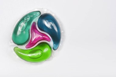 Tepede yeşil, mavi ve pembe sıvı bölümlü pervane şeklinde renkli bir deterjan kapsülü bulunur. Beyaz arka planda bolca fotokopi alanı bulunur.