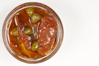 Yakın planda, cam bir kavanozda yağda muhafaza edilmiş güneşte kurutulmuş domatesler ve yeşil kapariler görülüyor.