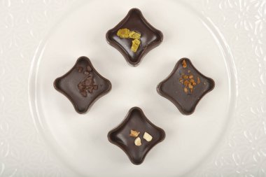 Üstten alta dört koyu çikolatalı pralin içerir, her biri eşsiz malzemelerle, beyaz kabartmalı bir tabakta ahşap bir yüzey üzerinde simetrik olarak sıralanmıştır.