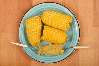  Üç sarı mısır koçanı ve mısır tutacağı ile yenmiş bir mısır koçanı yukarıdan ahşap bir yüzey üzerinde mavi bir tabakta sıralanır.