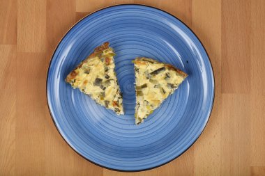  İki üçgen peynirli ve pırasalı kiş ahşap bir yüzeyde mavi bir seramik tabakta sunuluyor..