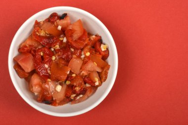 Kırmızı biberli, domatesli ve sarımsaklı canlı bir salata dolu beyaz bir kase çarpıcı bir kırmızı arka planda geniş bir kopya alanı ile yerleştirilmiştir.