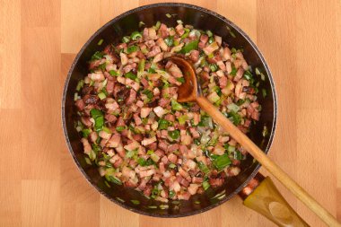 Yukarıdan bakıldığında, doğranmış pancetta ve doğranmış yeşil soğanlar, siyah bir tavada tahta bir kaşıkla karıştırılıyor.