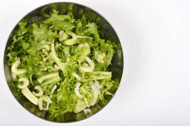 Bu yukarıdan aşağıya doğru, paslanmaz çelik kasede canlı yeşil bir salata sunar. Beyaz arka planda gevrek marul ve dilimlenmiş kereviz bulunur.