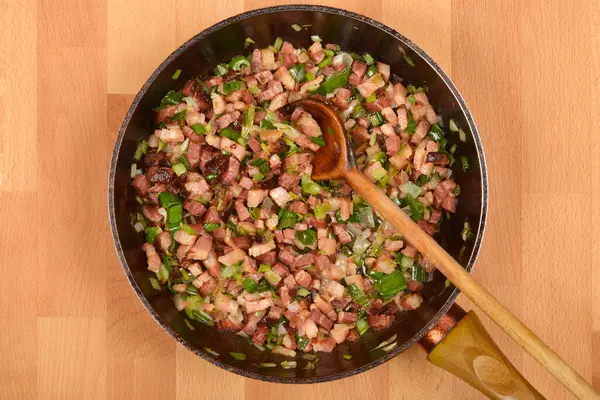 Yukarıdan bakıldığında, doğranmış pancetta ve doğranmış yeşil soğanlar, siyah bir tavada tahta bir kaşıkla karıştırılıyor.