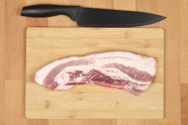 Ahşap bir kesme tahtasındaki çiğ domuz eti parçası. Üstünde siyah bir bıçak var. Yukarıdan aşağıya doğru çekilmiş.