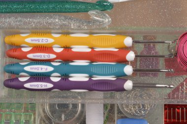 Dört ergonomik örgü kancasından oluşan renkli bir set, çeşitli el işi malzemeleriyle çevrili, temiz bir zanaat organizatöründe saklıdır.