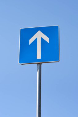 Mavi arkaplanı gösteren beyaz bir okla tek yönlü trafik işareti, açık mavi gökyüzüne karşı.