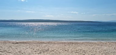 Selce 'deki kumlu Poli Mora plajından bir manzara, ön planda pırıl pırıl turkuaz deniz ve güneşli bir gökyüzünün altında Krk Adası.
