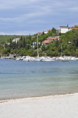 Jadranovo 'nun sakin kıyı manzarasında, kumlu bir sahil yakınındaki durgun turkuaz suya demirlemiş tekneler ve parçalı bulutlu bir gökyüzünün altındaki yeşil bir yamaçta yuva yapmış evler yer alıyor.