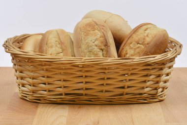 Kırsal bir dokuma sepetinin içi, sade beyaz bir arkaplana karşı ahşap bir masa üzerine kurulmuş taze pişmiş, altın-kahverengi ekmek ruloları ile doludur.