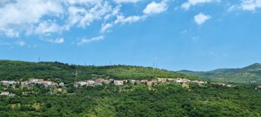 Bu resimde Praputnjak 'ın büyüleyici köyü, yemyeşil bir yamaçta, evleri ve yoğun ağaçların arasında, parçalı bulutlu bir gökyüzünün altında görülebilen bir kilisesi var.