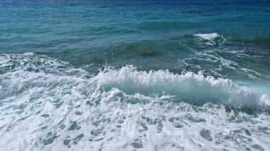 Güçlü bir dalga yükselir ve kırılır. Derin mavi ve turkuaz deniz boyunca uzanan canlı bir beyaz köpük çizgisi oluşturur.