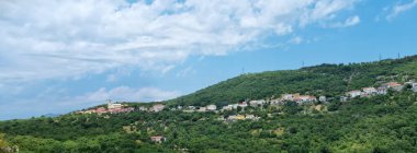 Bu resimde Praputnjak 'ın büyüleyici köyü, yemyeşil bir yamaçta, evleri ve yoğun ağaçların arasında, parçalı bulutlu bir gökyüzünün altında görülebilen bir kilisesi var.