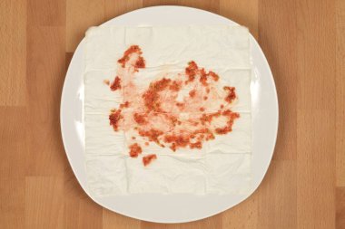 Beyaz bir tabakta, üzerinde kırmızı domates hamuru ve tohumlar olan kağıt bir peçete bulunur. Ahşap bir arka plana kurulanır.