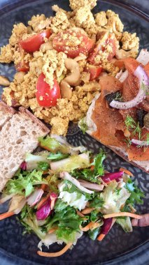 Canlı bir vejetaryen kahvaltı tabağında domatesli yumuşacık tofu, bagel üzerine havuç somonu, taze salata ve dilimlenmiş ekmek yer alır.