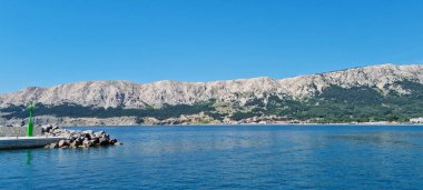 Krk Adası 'ndaki Baska' daki yeşil rıhtımda sakin bir deniz var. Uzakta Zarok ve Vela Plaza heybetli bir dağ sırasının altında görünüyor.
