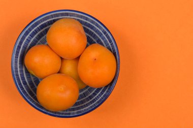 Beş olgun, turuncu kayısıyla dolu küçük, mavi ve beyaz bir kase canlı turuncu bir zemin üzerine yerleştirilmiş, geniş bir kopya alanı var.