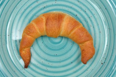 Altın-kahverengi, pullu bir kruvasan, merkezi olarak eşsiz bir turkuaz tabağa yerleştirilir. Konsantrik çıkıntılı, iştah açıcı dokusunu ön plana çıkarır.