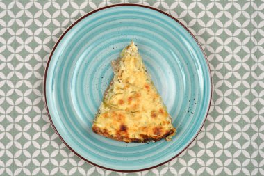 Bir dilim baharatlı kabak ve pırasalı kiş kahverengi kenarlı mavi bir seramik tabakta desenli bir masa örtüsüne karşı sergilenir.