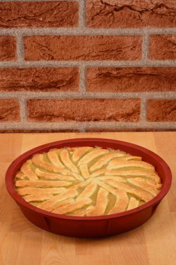 Dekoratif kabuklu yeni pişmiş elmalı turta ahşap bir yüzey üzerinde kırmızı silikon bir pişirme kabında sunuluyor ve arka planda tuğla bir duvar bulunuyor.