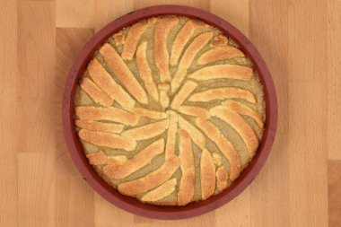 Tepeden çekilen bir fotoğraf, hafif ahşap bir yüzey üzerinde dinlenen, kırmızı bir fırın kabuğunda dekoratif bir kabukla taze pişmiş bir elmalı turta yakalar.