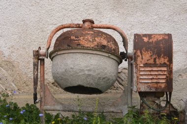 Eski, paslı bir beton mikser duvarın önünde duruyor, etrafını yabani otlar ve yabani çiçekler sarmış.