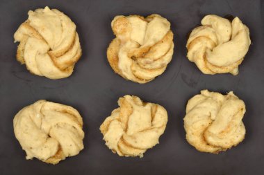 An overhead studio shot displays six unbaked cardamom rolls (kardemummabullar) shaped in swirls on a dark baking sheet