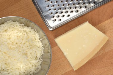 Tepeden çekimde parmesan peyniri, metal kasede rendelenmiş peynir ve tahta kesme tahtasındaki peynir rendesi görülüyor.