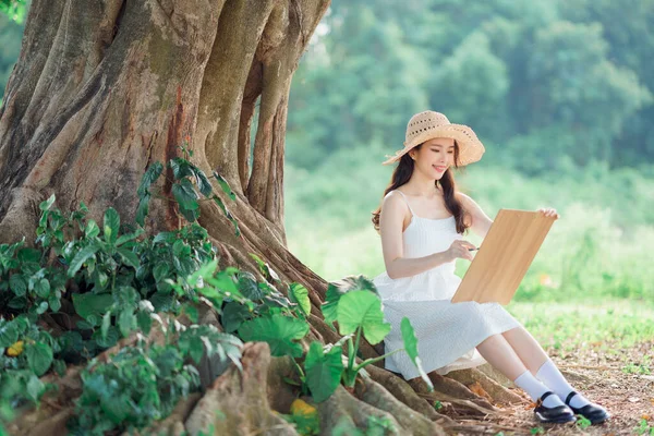 The girl is drawing under the big tree