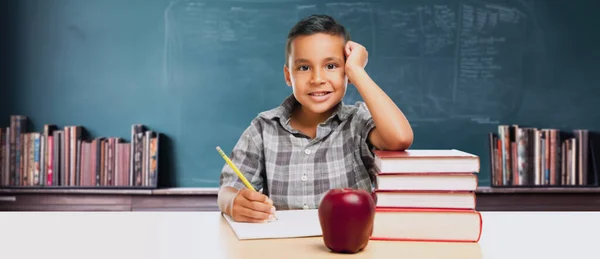 チョークボードの前で本とアップルが付いているデスクで幸せな若いヒスパニックの学校の少年 — ストック写真